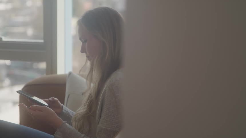 A woman with long blonde hair wearing a light gray sweater sits on a couch and uses a mobile phone with a bright window visible in the background