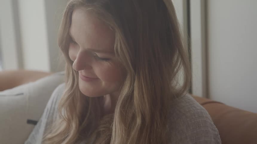 A woman sits on a couch in the living room while looking down at something and smiling