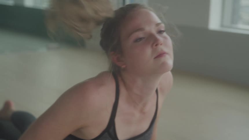 A young woman with blonde hair in a ponytail stretches on the floor of an apartment with large windows