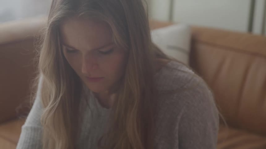 A young woman with long blonde hair looks down with a pensive expression her face partially obscured by a soft blur against the backdrop of a couch