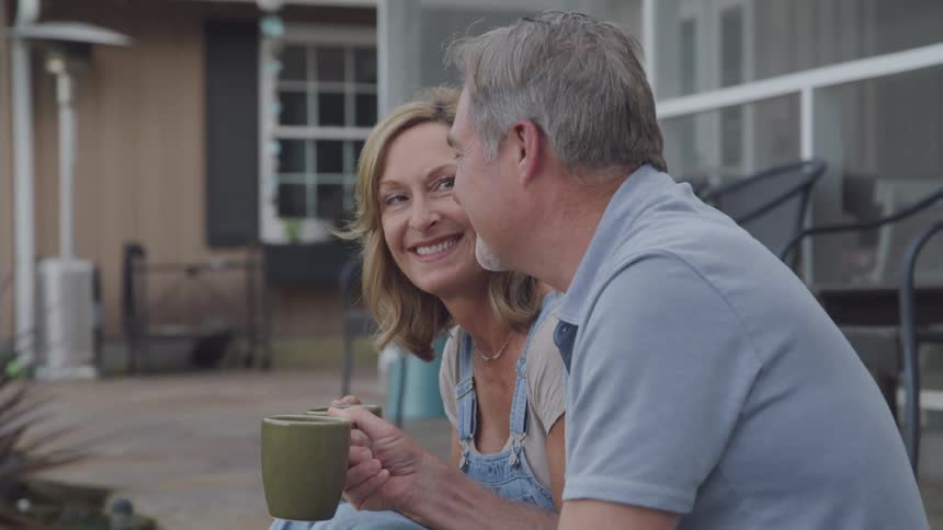 A smiling elderly couple sits outdoors enjoying a quiet moment together while holding mugs