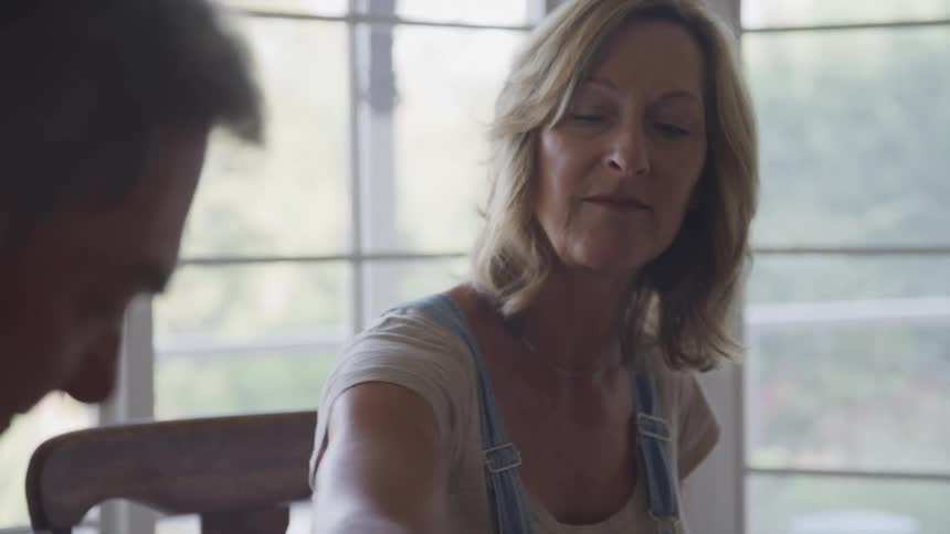 Two people sit at a table in front of a large window overlooking trees the woman wearing a lightcolored shirt and denim overalls