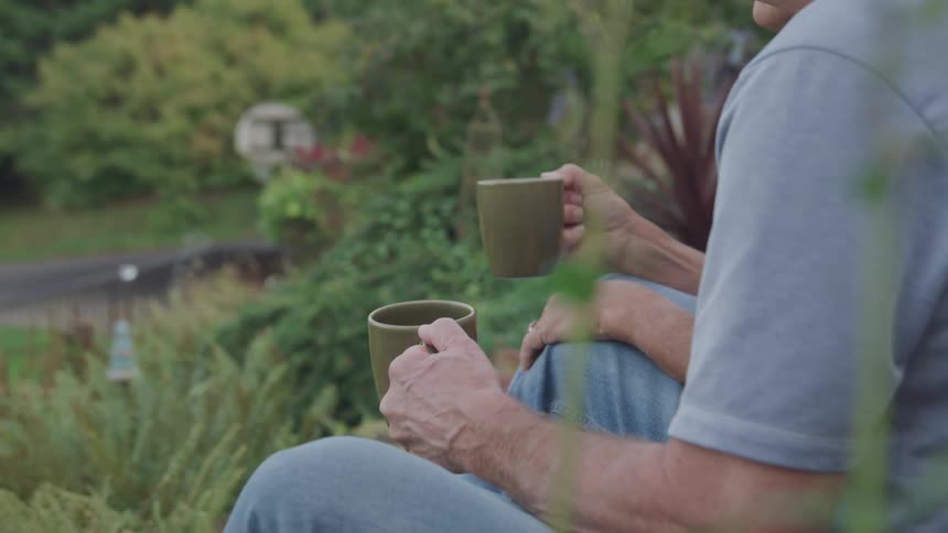 Couple sitting outdoors overlooking their garden holding coffee mugs