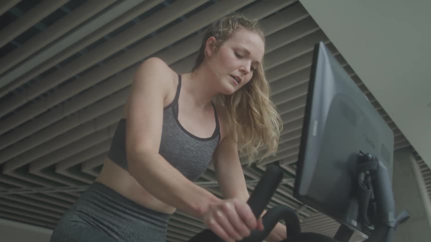 A woman with blonde hair pulled back in a ponytail works out on a stationary bike in a gym wearing a sports bra and leggings with a focused expression and a computer screen visible in the foreground