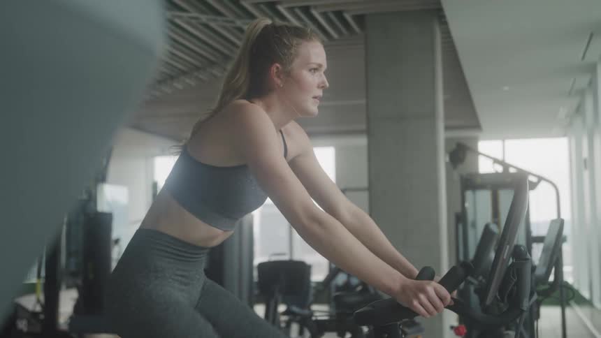 A woman with blonde hair pulled back in a ponytail works out on a stationary bike in a gym wearing a sports bra and leggings with a focused expression