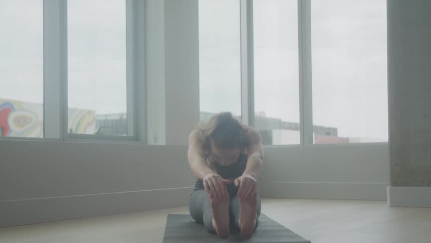 A woman with dark hair pulled back in a ponytail sits on a yoga mat stretching her arms forward in a seated forward bend with a large window and cityscape visible in the background
