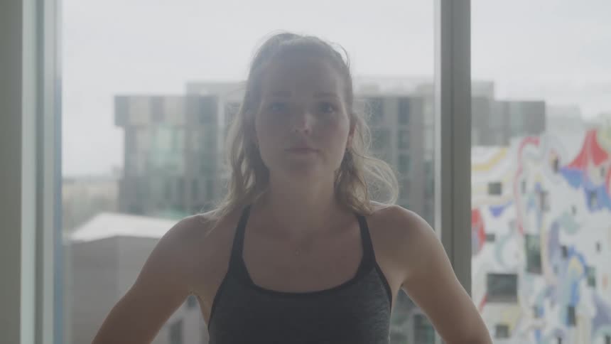A woman with blonde hair pulled back in a ponytail stands facing the camera wearing a sports bra with a bright window and cityscape visible in the background