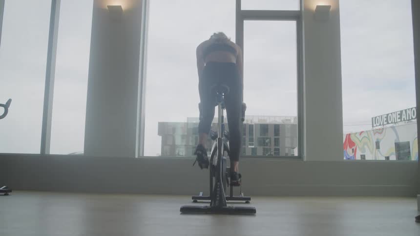 A woman with blonde hair pulled back in a ponytail works out on a stationary bike in a gym wearing a sports bra and leggings with a focused expression and a large window overlooking a cityscape