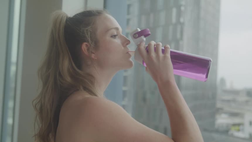 A woman with long blonde hair wearing a sports bra drinks from a pink water bottle against the backdrop of a city skyline seen through a window