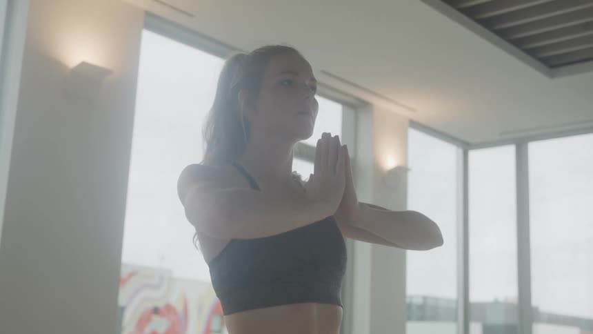 A woman with dark hair pulled back in a ponytail stands in a yoga pose with her hands in prayer position wearing a sports bra with a bright window visible in the background