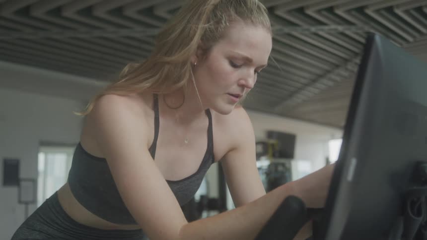 A woman with blonde hair pulled back in a ponytail works out on a stationary bike in a gym wearing a sports bra and leggings with a focused expression