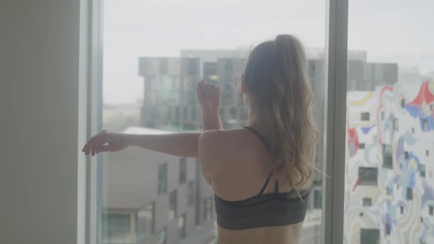 A medium shot shows a woman in athletic wear stretching by a window with a cityscape visible beyond