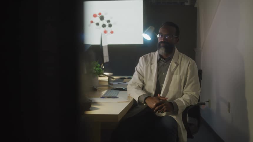 A person in a lab coat sits at a desk surrounded by computer monitors a lamp and scientific equipment with a molecular model displayed on a screen in the background