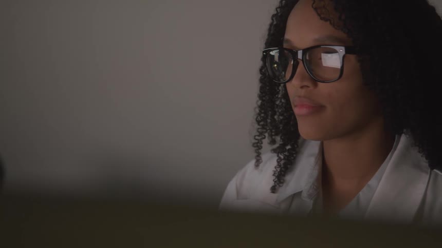 A young woman with curly black hair and thick glasses works at a computer in a dimly lit office