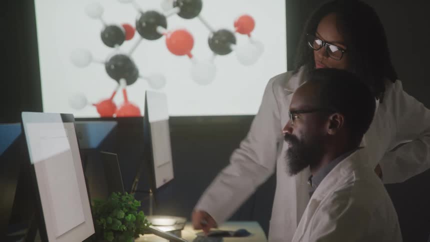 A man and a woman in white lab coats look at a computer screen