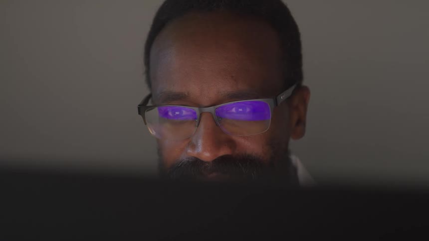 A doctor with dark hair a beard and glasses looking intently at a laptop screen in a dimly lit environment
