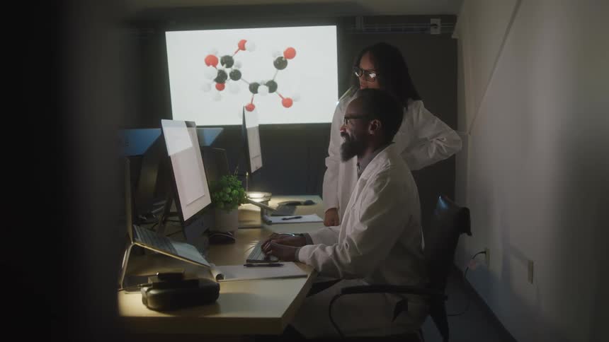 Two individuals in lab coats work at computer stations in a dimly lit room with a molecular structure projected on a screen in the background