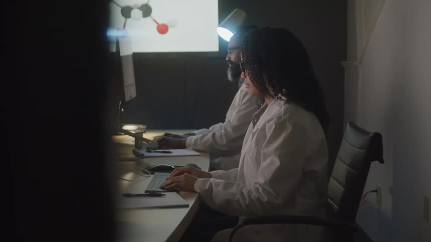 Two individuals possibly scientists wearing lab coats work at separate computer workstations in a dimly lit room looking at screens displaying scientific visuals
