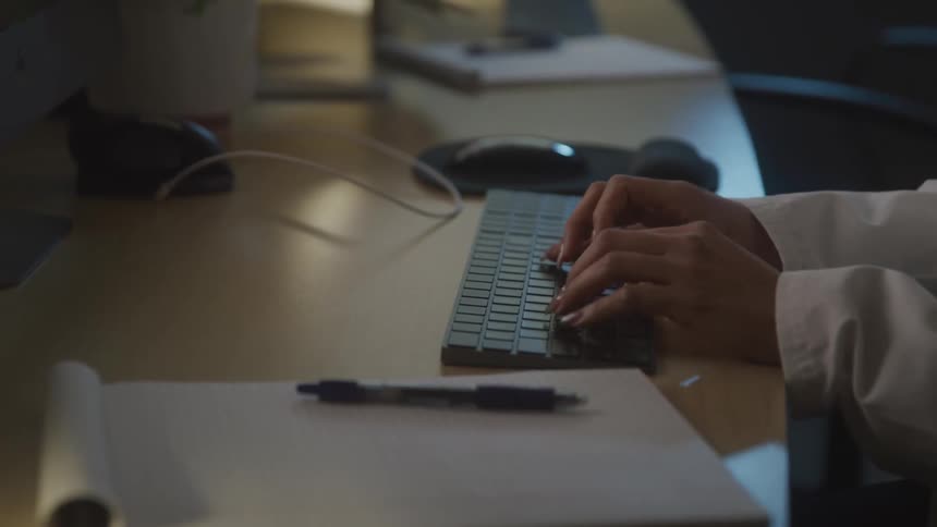 Hands typing on a keyboard with a pen and notepad in the foreground working in a dimly lit setting