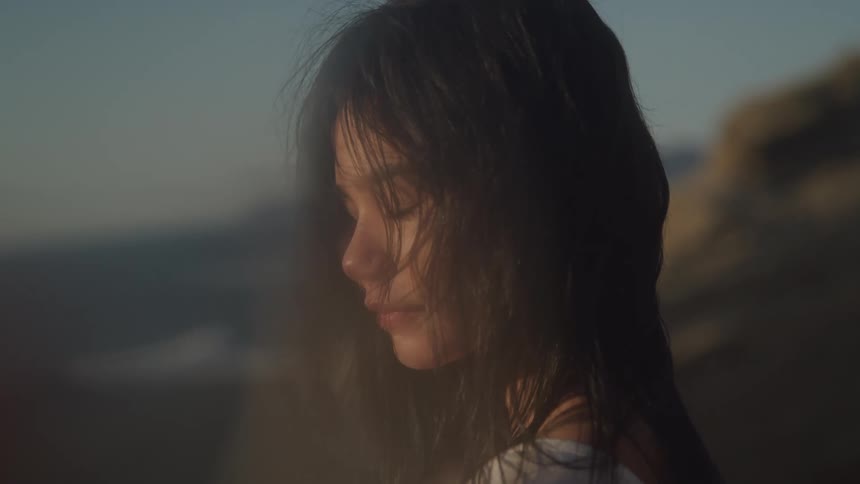 A young woman with dark tousled hair looks down with a thoughtful expression her face partially obscured by a soft blur and the outdoor setting