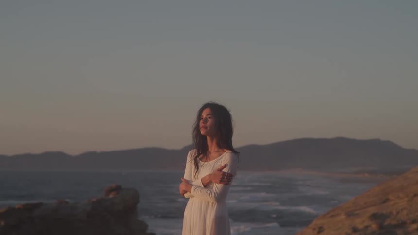 A woman in a dress stands between two giant rocks with a mountain range behind her