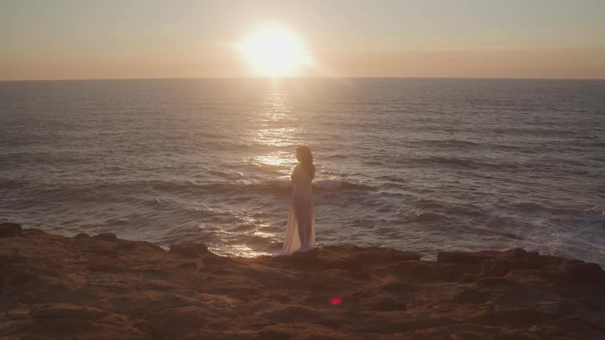 Woman wearing a translucent skirt stands on the coast while the sun sits on the horizon