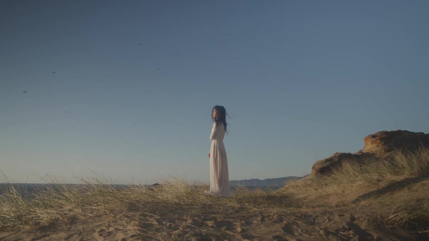A woman in a white dress stands on a grassy knoll while looking into the distance