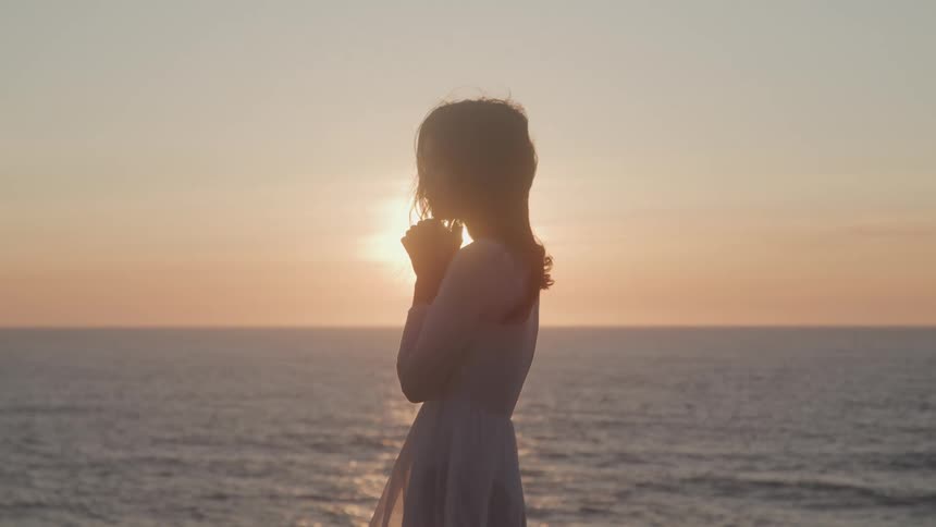 A woman in a white dress stands hands clasped near the water at sunset