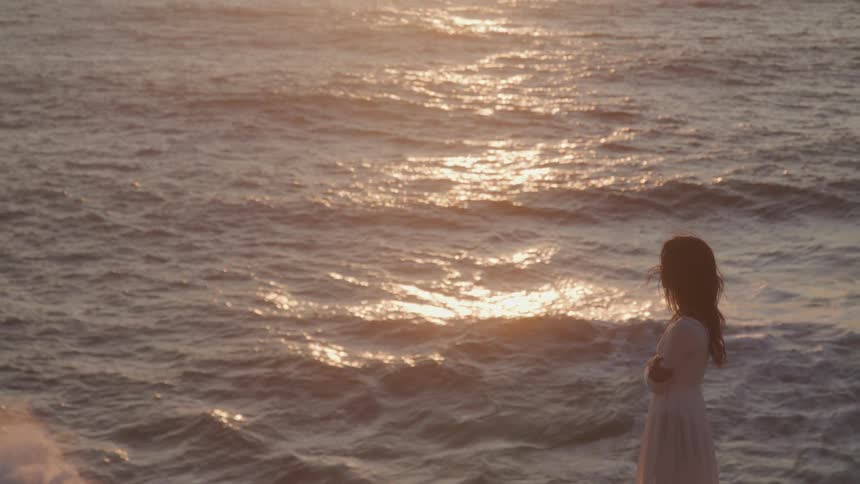 A woman in a light colored dress stands in silhouette against the backdrop of a shimmering sunlit ocean at sunset