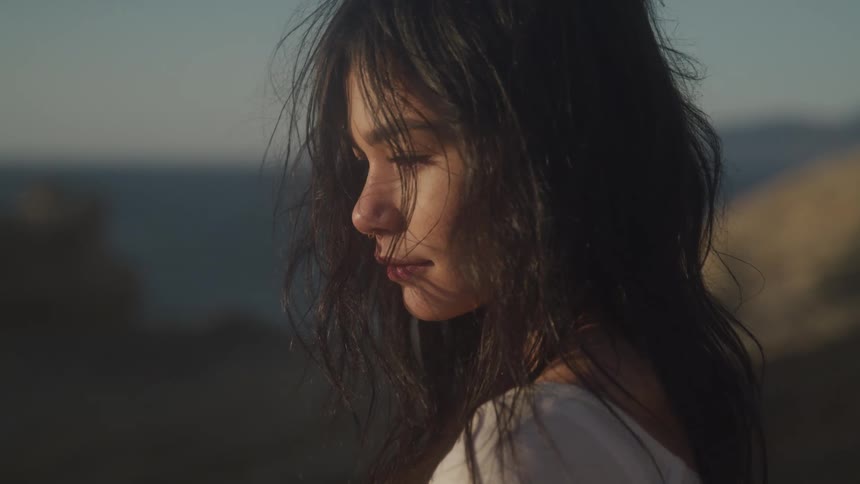 A young woman with dark tousled hair her face illuminated by soft light as she looks down with a contemplative expression