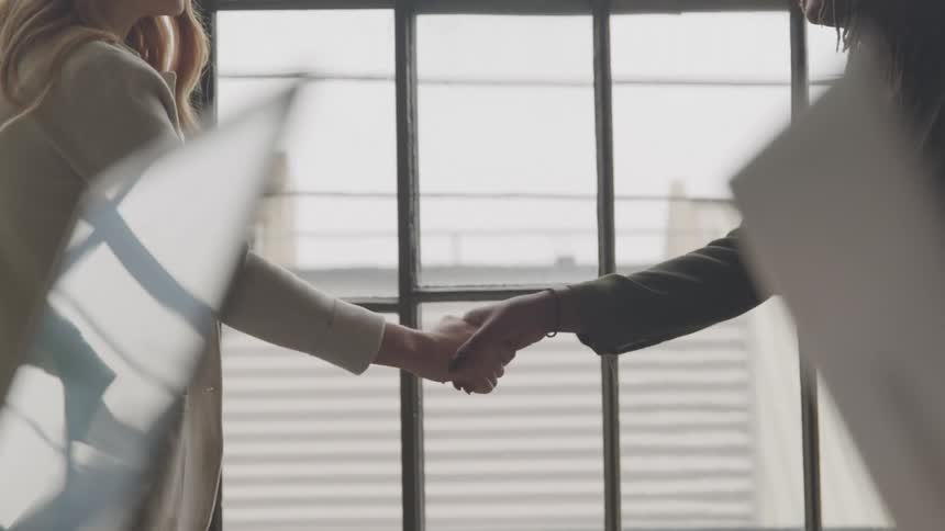 Two professional females shaking hands in an office setting in front of a window