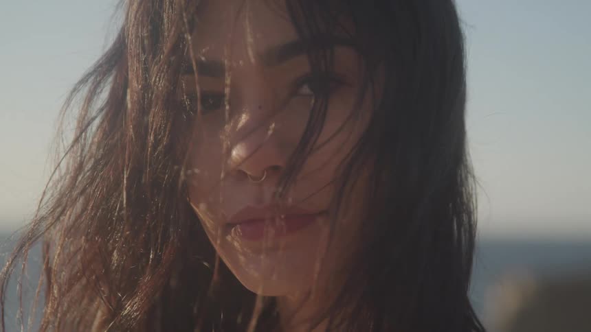 A young woman with dark tousled hair framing her face with a hint of a nose ring visible against a blurred outdoor background