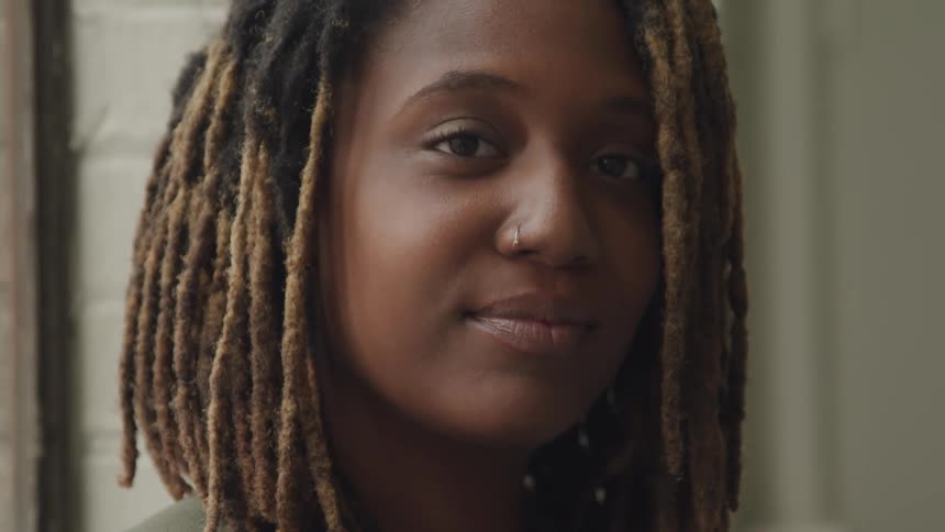 A woman with dreadlocks who is looking slightly to the side with a gentle smile against a soft neutral backdrop
