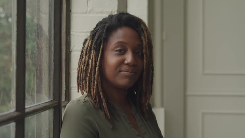 A woman with dreadlocks looks directly at the camera standing near a window with foliage visible outside