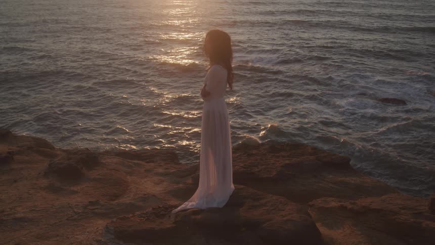 A woman in a flowing white dress stands on a rocky cliff overlooking the ocean at sunset