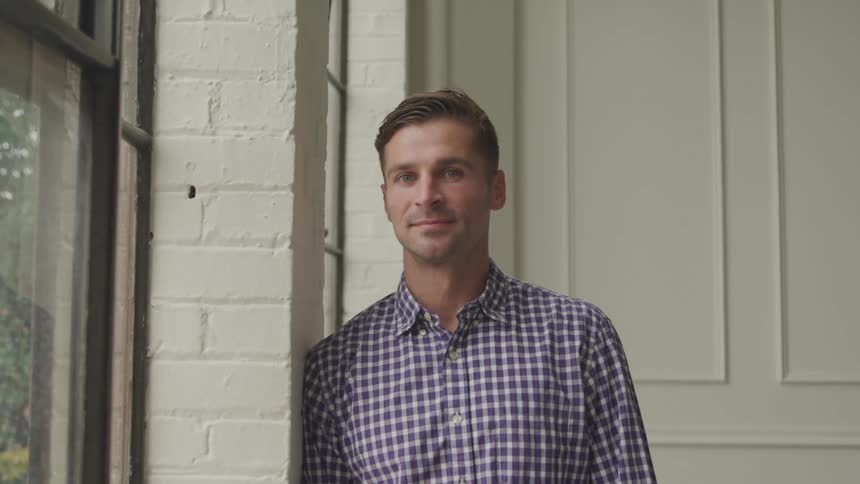A man with light brown hair wearing a blue and white checkered shirt leans casually against a wall next to a window looking directly at the camera with a slight smile