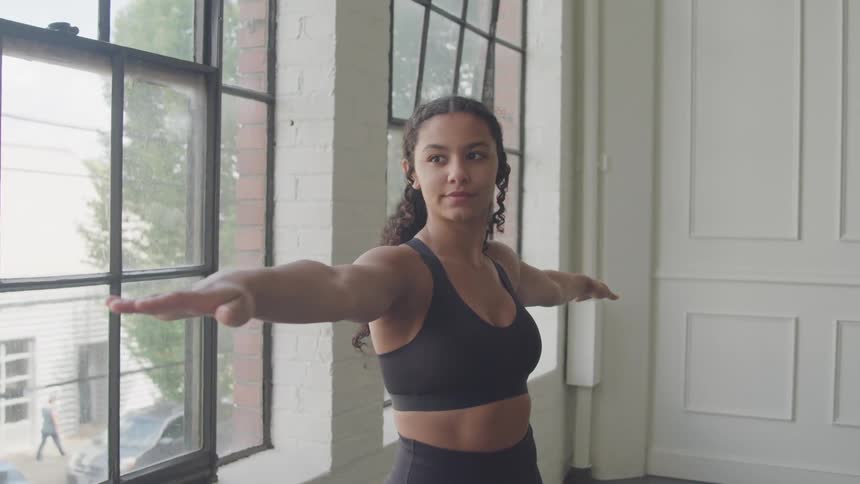 A young woman with curly dark hair wearing a black sports bra and leggings is captured midyoga pose with her arms outstretched looking slightly away from the camera in a room with large windows and white paneled walls