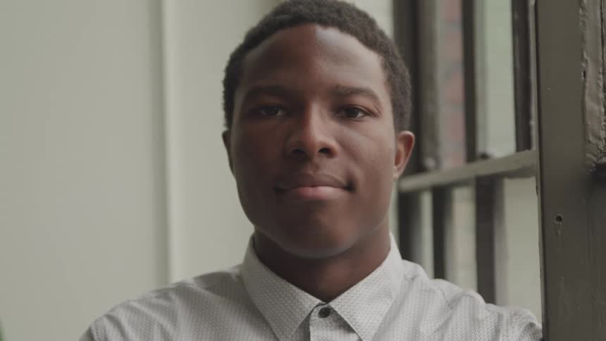 A young man with short dark hair wearing a light colored button down shirt looks directly at the camera with a slight smile