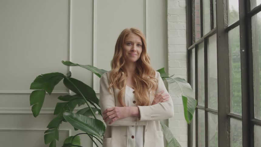 A woman with long reddish blonde hair stands with her arms crossed smiling slightly in a room with large windows and a potted plant