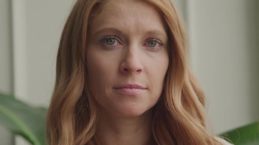 A woman with long wavy red hair looks directly at the camera with a neutral expression