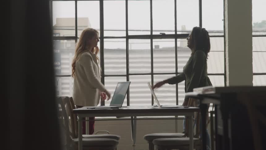 Two women in formal attire shake hands across a table with laptops in an office featuring large windows