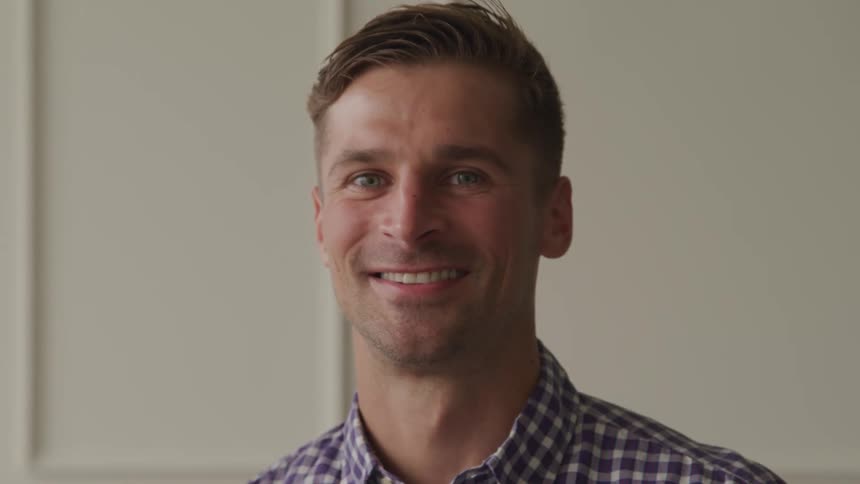 A smiling man in a plaid shirt poses against a white wall looking directly at the camera