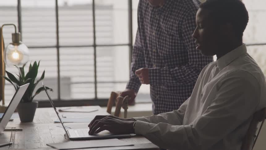 Two individuals collaborate at a desk with a laptop papers and a potted plant one standing and gesturing at the screen while the other types