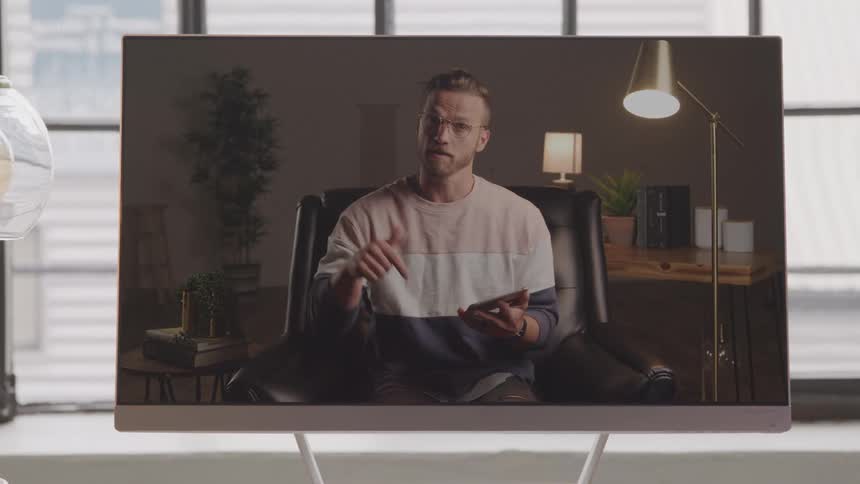 A man appears on a large monitor seemingly engaged in a video call or presentation within a well lit modern setting