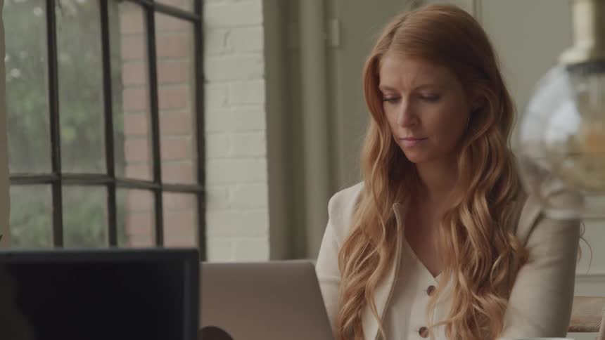 A woman with long wavy red hair sits at a desk looking down with a pensive expression working on a laptop in a room with a large window and a partially visible lamp