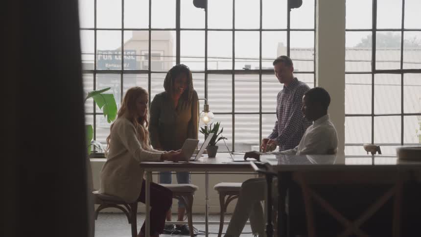 Four diverse colleagues engage in a meeting at a bright modern office space illuminated by natural light streaming through expansive windows