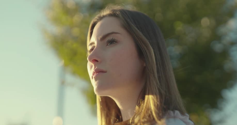 A young woman with long straight brown hair looks off into the distance with a thoughtful expression her face illuminated by soft sunlight
