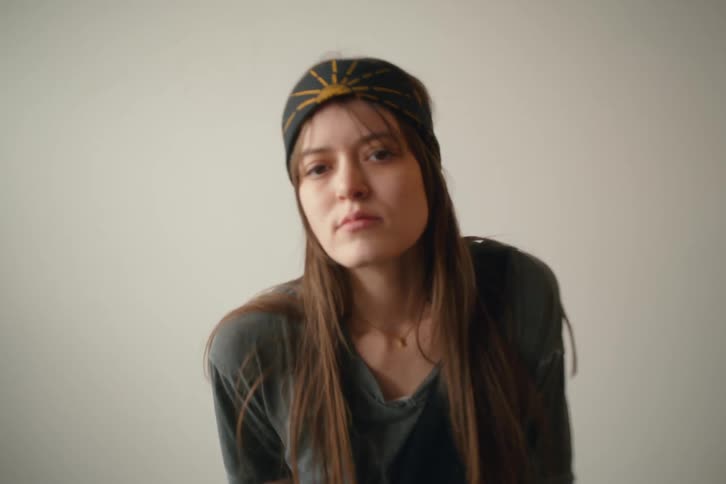 A young woman with long brown hair wearing a patterned headband looks directly at the camera with a neutral expression against a plain light colored background