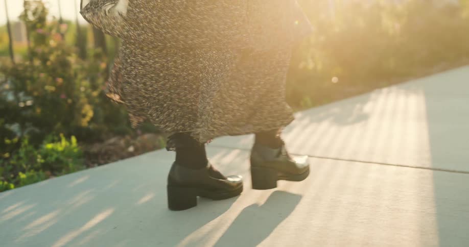 A person wearing a patterned skirt and black heeled boots walks on a concrete surface bathed in warm sunlight