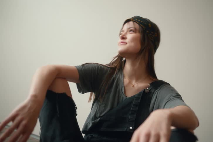 A young woman with long brown hair wearing a gray tshirt black overalls and a patterned beanie sits casually on the floor smiling and gesturing with her hands